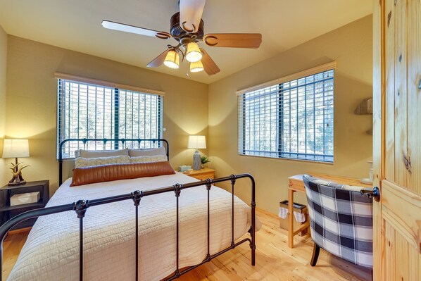 A bedroom with a bed, two bedside tables, a chair, a ceiling fan, and two windows with blinds.
