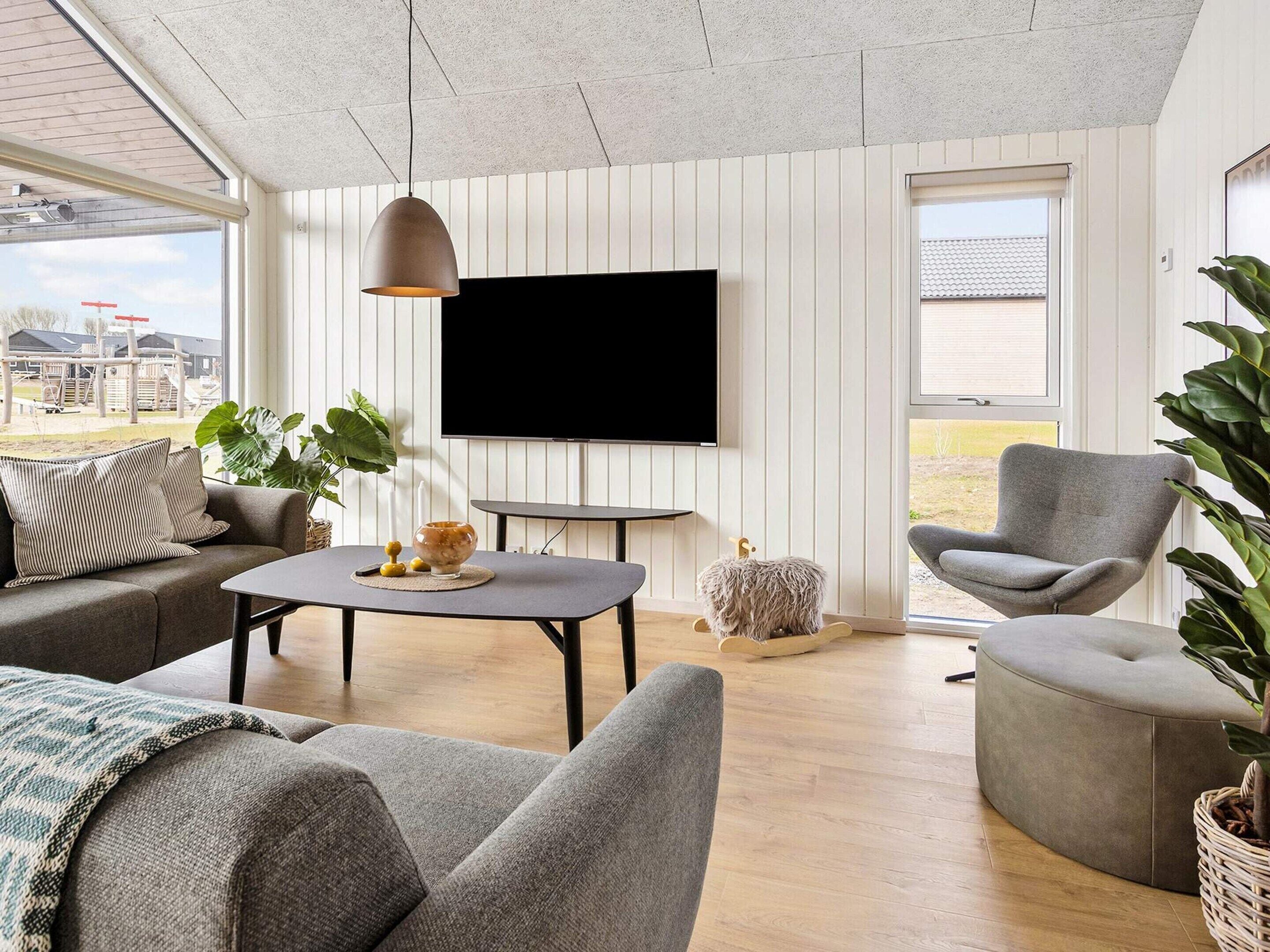 Una sala de estar moderna con sofá, sillón y una mesa con un bol y velas. Un televisor está montado en la pared, y hay una ventana grande con vistas al exterior.