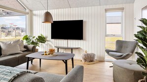 Een moderne woonkamer met een bank, fauteuil en een tafel met een schaal en kaarsen. Een televisie is aan de muur bevestigd, en er is een groot raam met uitzicht op buiten.