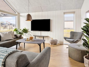 Un salon moderne avec un canapé, un fauteuil et une table sur laquelle se trouvent un bol et des bougies. Un téléviseur est fixé au mur, et une grande fenêtre offre une vue sur l’extérieur.