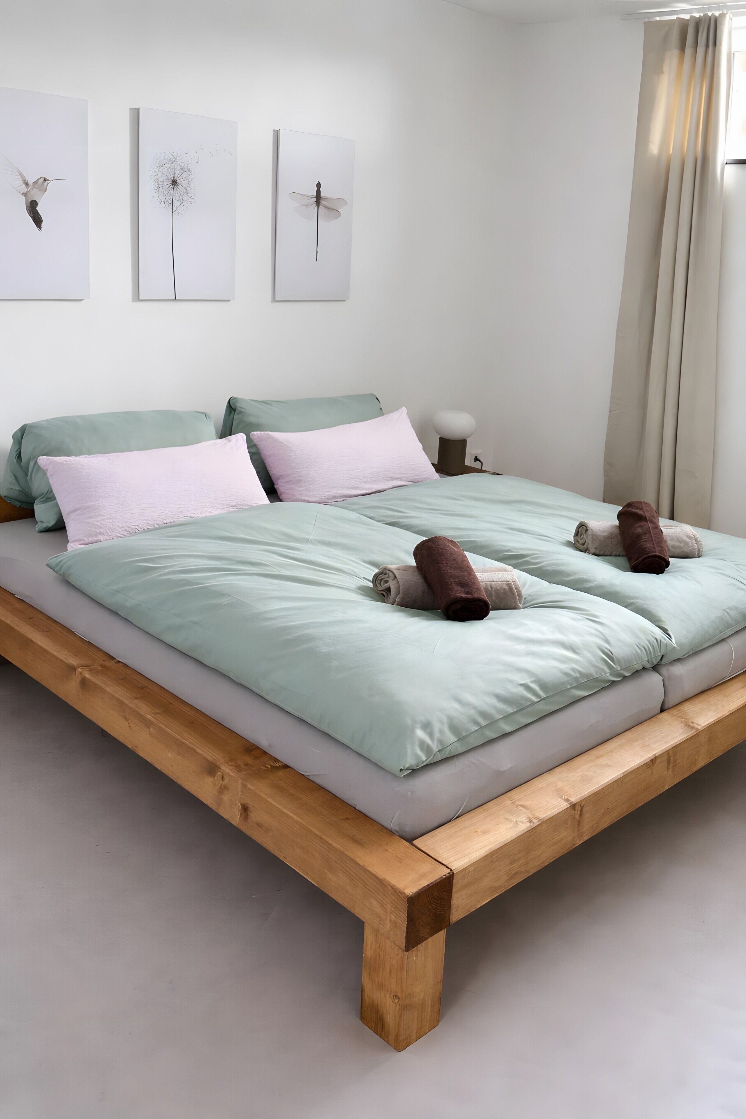 Una cama con estructura de madera, ropa de cama verde claro y dos toallas enrolladas.