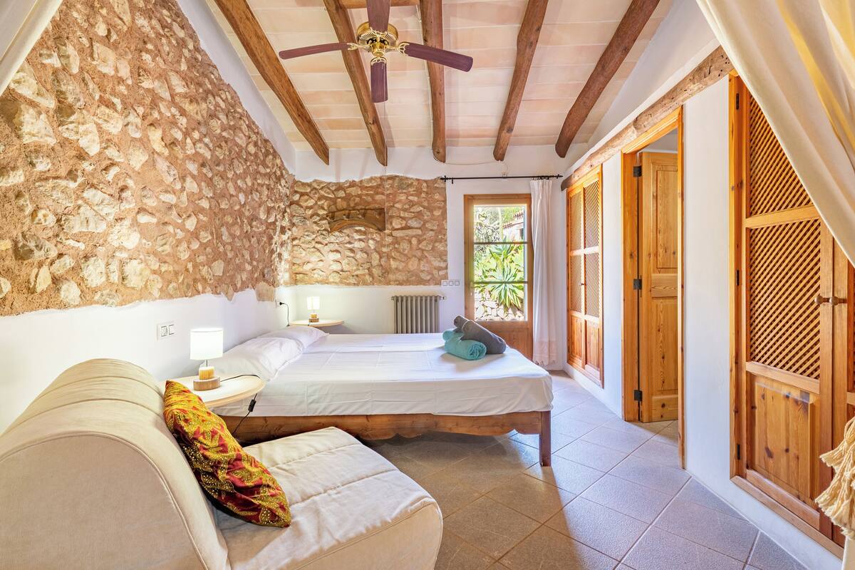 Une chambre avec un mur en pierre, des poutres en bois, un lit, une table de chevet, une lampe et une fenêtre donnant sur un paysage verdoyant.
