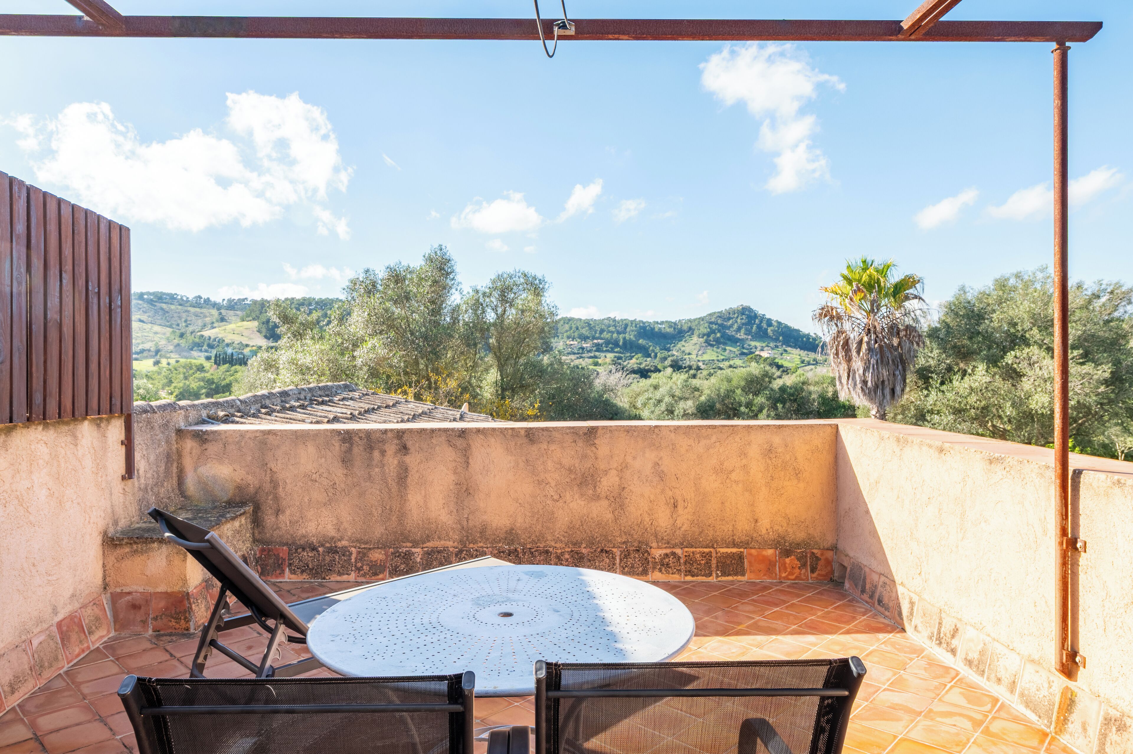 A terrace with a round table and chairs, overlooking a hilly landscape with trees and a palm tree.