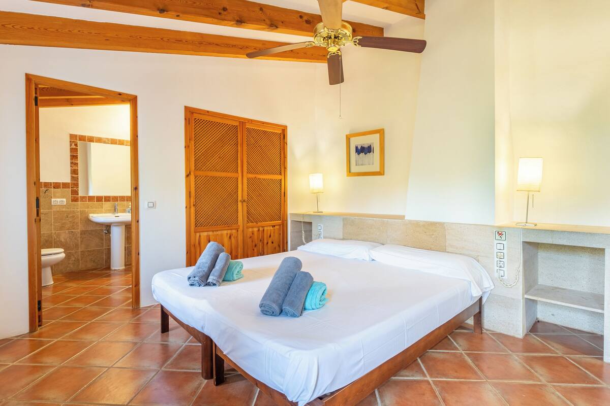 Une chambre d’hôtel comprenant un lit, deux serviettes, un ventilateur de plafond, une salle de bain avec un lavabo et des toilettes, et une porte en bois.