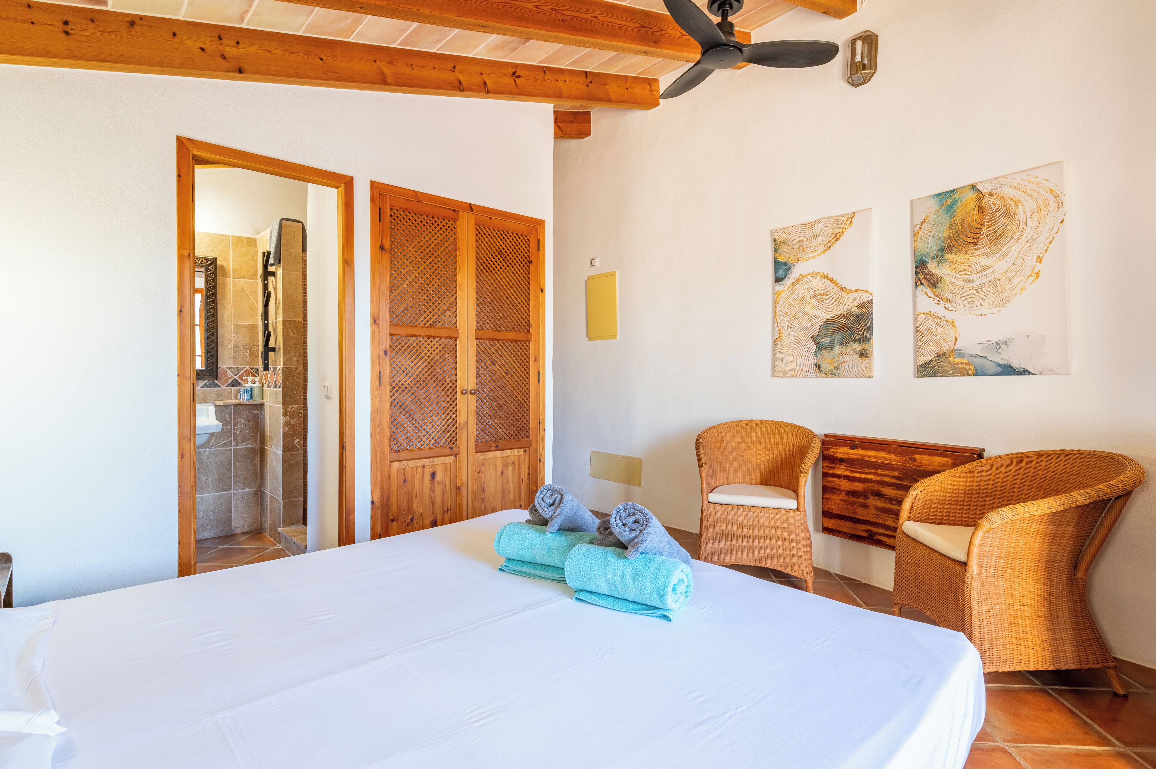 A hotel room with a bed, two wicker chairs, a ceiling fan, and artwork on the wall.