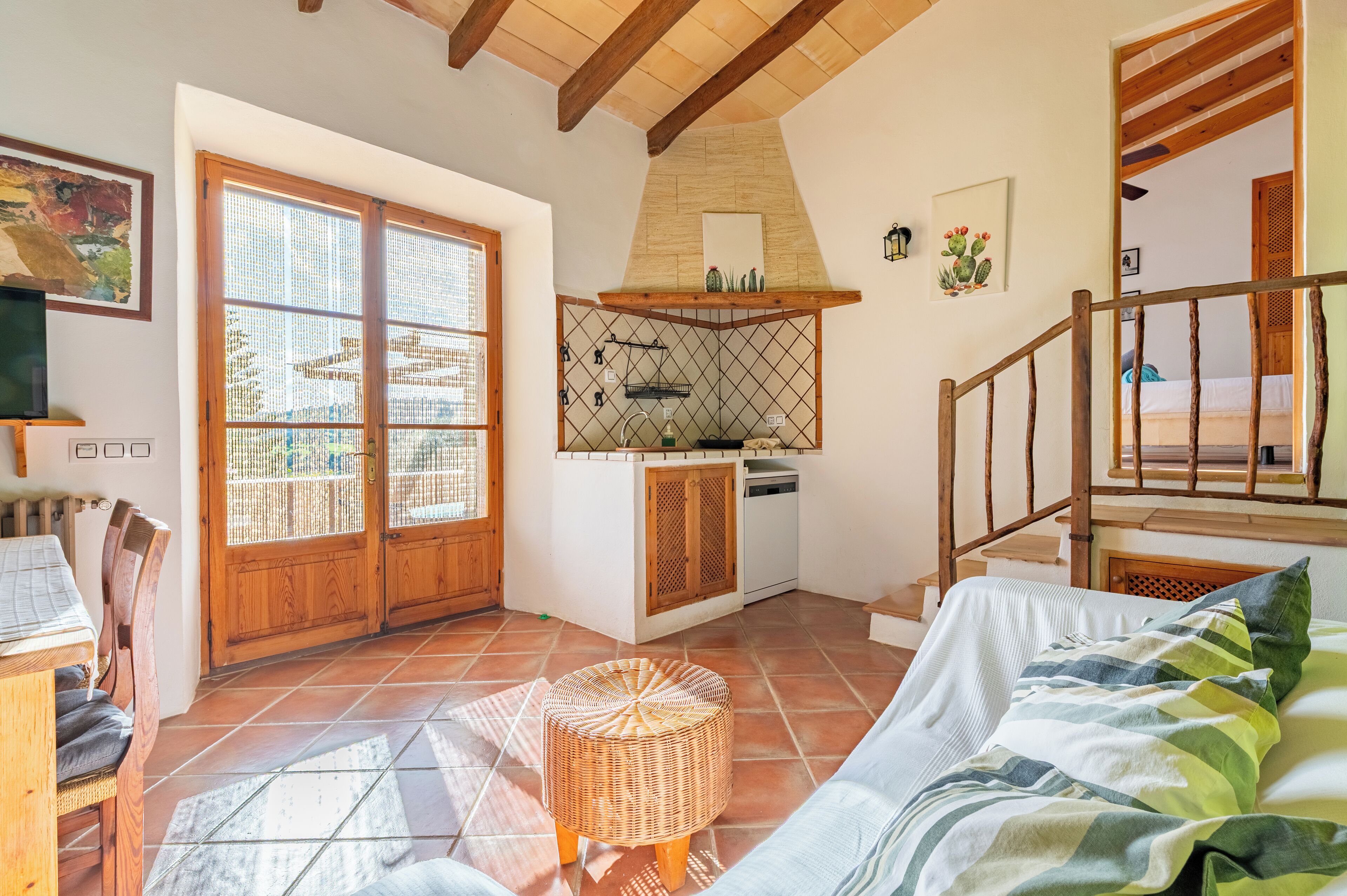 A cozy room with a wooden ceiling, a kitchen area with a hanging pot rack, a bed with a striped blanket, and a wicker stool.