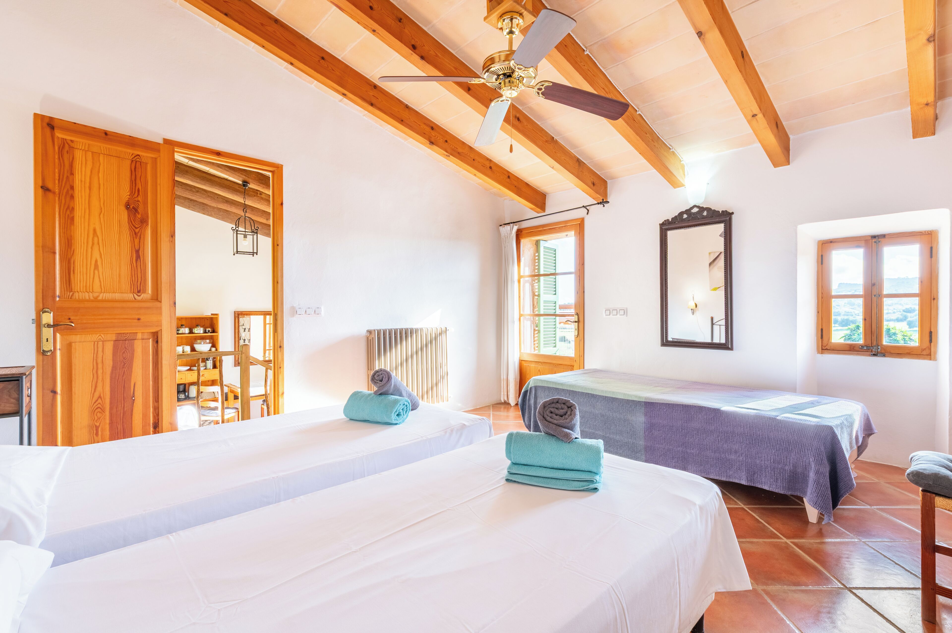 A room with two beds, wooden beams, a ceiling fan, a radiator, and a window with shutters.