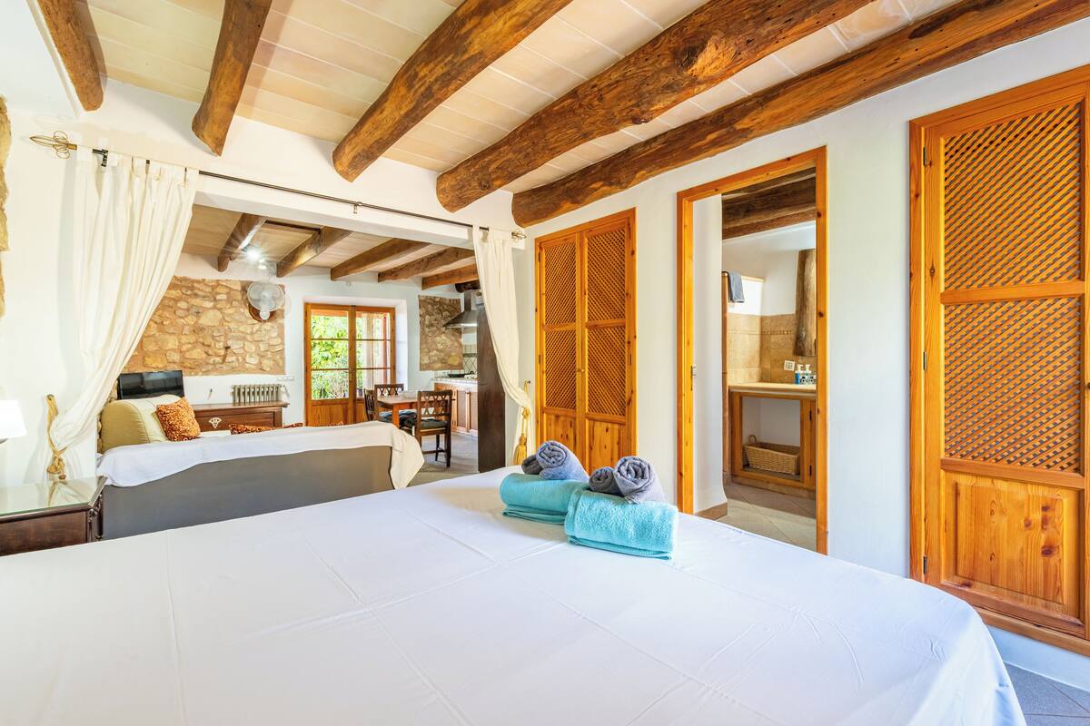 Une chambre spacieuse avec un grand lit, des poutres en bois, un mur en pierre et une vue sur l’espace repas.