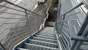 Escadaria de metal que leva à entrada de um prédio, com um SUV branco estacionado na frente.