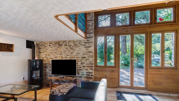 A living room with a stone wall, a sofa, a glass coffee table, a TV, and a wooden staircase. - Charming Cottage in the Périgord Vert Countryside (Angoisse)