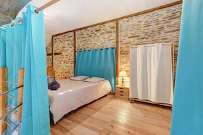 A bedroom with a stone wall, a bed with a patterned bedspread, a nightstand with a lamp, and a wooden floor. - Charming Cottage in the Périgord Vert Countryside (Angoisse)