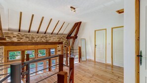 A spacious loft bedroom with a sloped ceiling, wooden beams, a stone wall, and a view through large windows. - Charming Cottage in the Périgord Vert Countryside (Angoisse)
