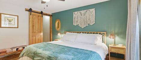 A bedroom with a bed, a ceiling fan, a wall-mounted macrame piece, a framed picture, and a wooden door.