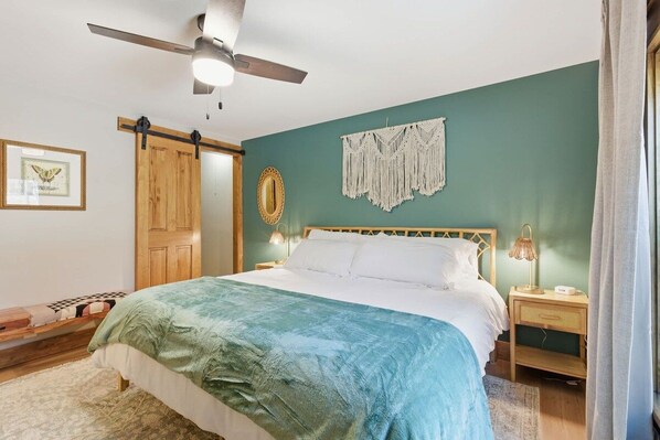 A bedroom with a bed, a ceiling fan, a wall-mounted macrame piece, a framed picture, and a wooden door.
