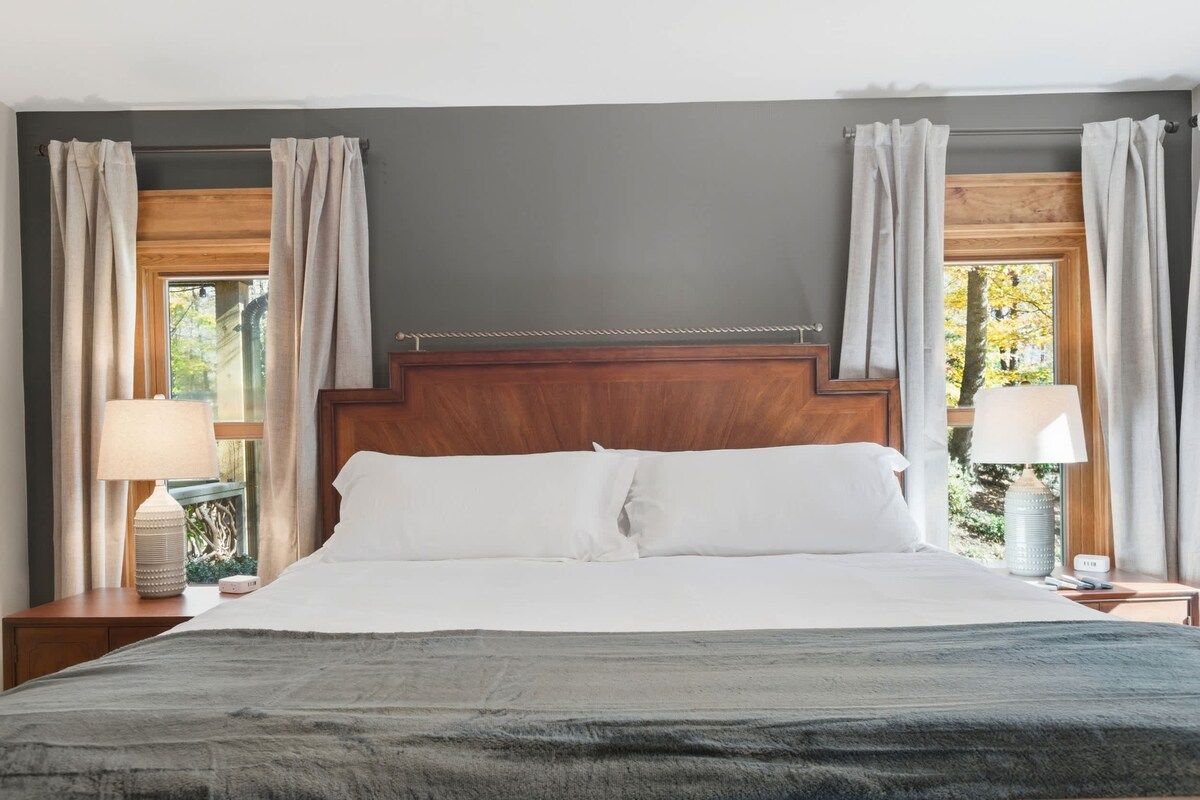 Une chambre à coucher avec un grand lit, une tête de lit en bois, deux tables de chevet avec des lampes et une fenêtre avec des rideaux.