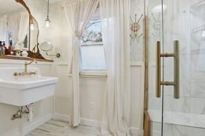A bathroom with a white sink, a mirror with a wooden frame, a shower with a glass door, and a window with curtains. - Cute Cottage, Central Location, Pet Friendly (Greer)