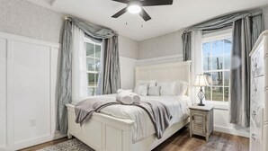 A bedroom with a white bed, a ceiling fan, a nightstand with a lamp, and a view of the outdoors through a window.