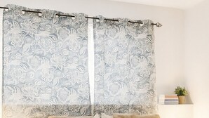 Quarto com uma cama grande, duas mesas de cabeceira, um criado-mudo, uma luminária, uma planta em vaso e uma janela com cortinas estampadas.