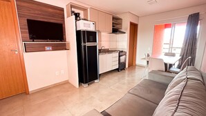 A modern kitchen with a black refrigerator, a microwave, and a stove. There is a dining area with a table and chairs, and a living room with a sofa. - Apto com Wi Fi Garagem e Piscina a 100m da Praia (Luis Correia)