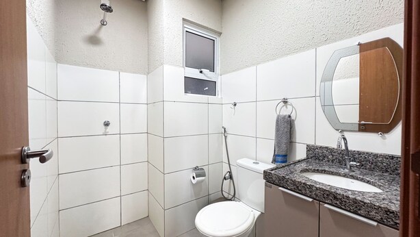 A bathroom with white tiles, a shower, a toilet, a sink with a granite countertop, and a mirror. - Apto com Wi Fi Garagem e Piscina a 100m da Praia (Luis Correia)