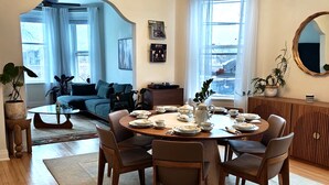 A dining area with a round table set for four, surrounded by chairs. There is a sofa and a coffee table in the background, and a large window with curtains. - Logan Sq 4BR Duplex • Garage • Sleeps 8 (Chicago)