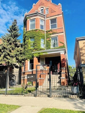 A two-story brick building with a prominent corner entrance, a gated driveway, and a large tree in front. - Logan Sq 4BR Duplex • Garage • Sleeps 8 (Chicago)