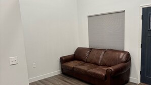 Una habitación con sofá de cuero marrón, ventana con persianas y suelo de madera oscura.