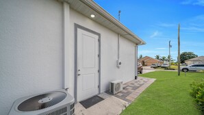 Un edificio residencial con fachada blanca, una puerta principal, un equipo de aire acondicionado, un pequeño patio empedrado y un césped.