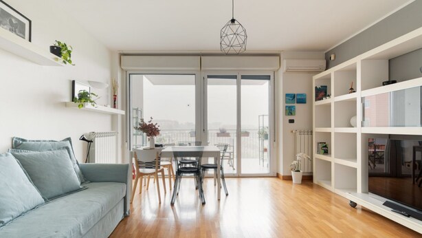 Un salon moderne comprenant un canapé, une table à manger, des chaises, une bibliothèque blanche et une vue sur l’extérieur, visible à travers une porte vitrée.