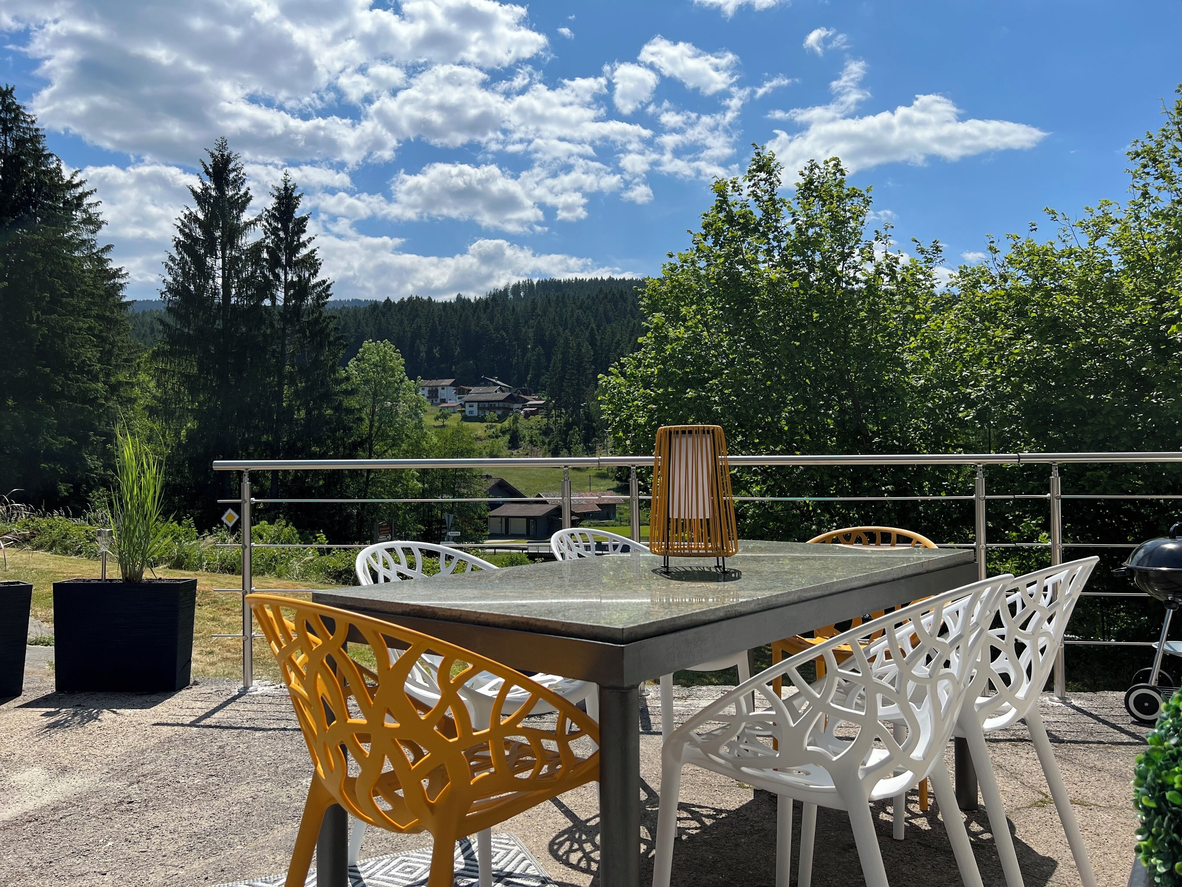 Une terrasse extérieure avec une table et des chaises, un barbecue et une vue sur une zone boisée, avec des maisons en arrière-plan.