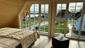 Ein Schlafzimmer mit einem Bett, einem schwarzen Hocker und großen Fenstern mit Blick auf eine Wohngegend.