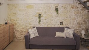 A cozy room with a stone wall, a wooden ceiling, a grey sofa with patterned cushions, a small table with a lamp, and a wooden cabinet. - Vacation home La Ficurinia in the heart of the historic center of Syracuse (Siracusa)