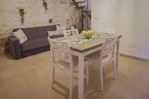 Un espace repas comprenant une table en bois, des chaises blanches, un canapé gris et un mur en pierre.