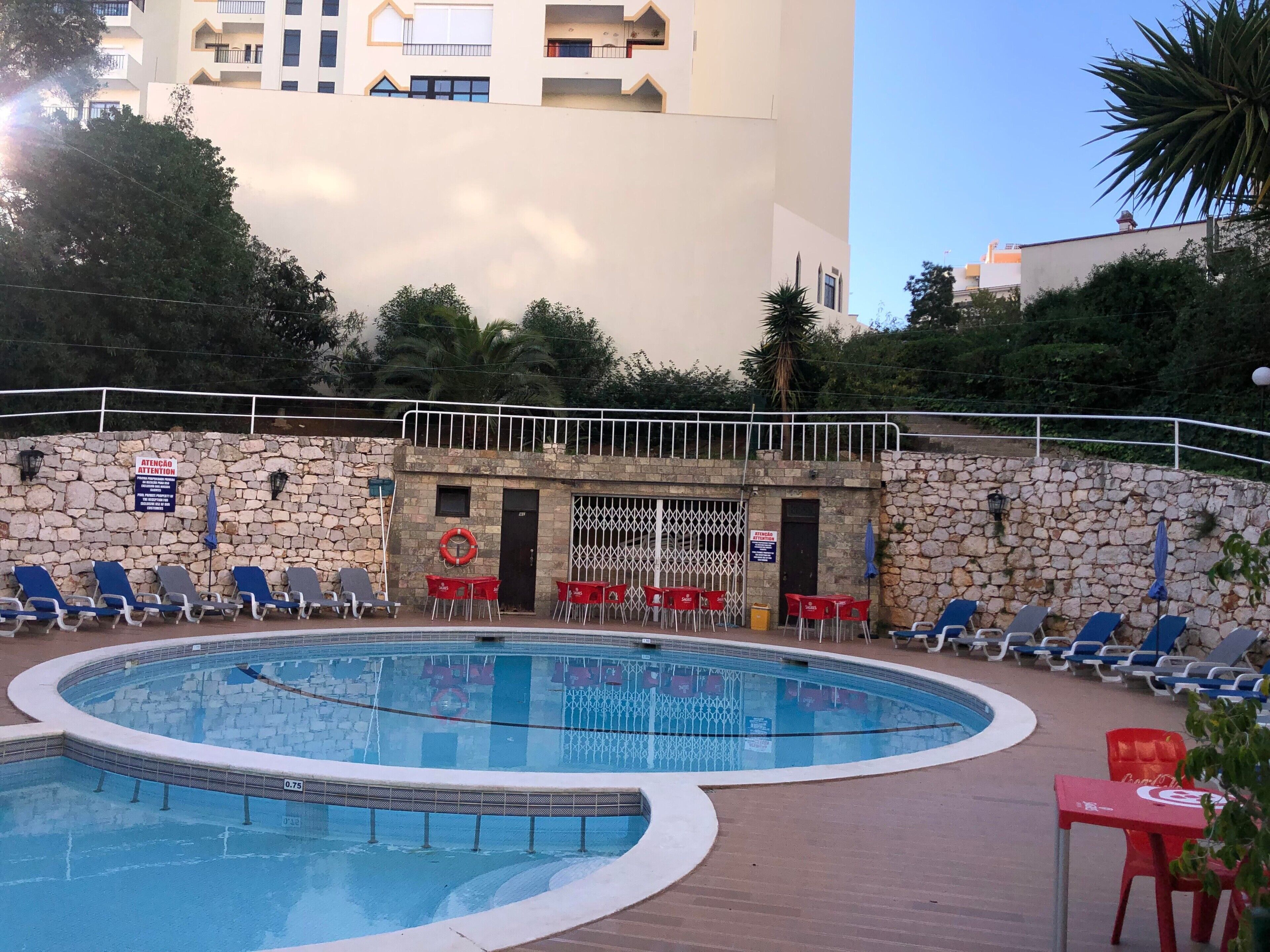 Una piscina con sillas de descanso, un muro de piedra y un edificio al fondo.