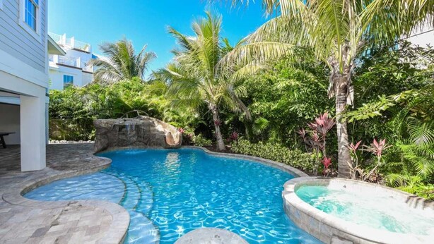 A swimming pool with a stone edge, surrounded by lush tropical vegetation and a white building in the background. - Malibu Beach ~ Waterslide/Rooftop Views/Walk to Beach (Holmes Beach)