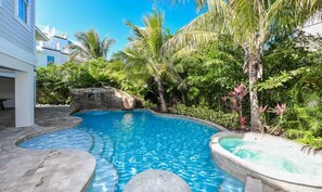 A swimming pool with a stone edge, surrounded by lush tropical vegetation and a white building in the background. - Malibu Beach ~ Waterslide/Rooftop Views/Walk to Beach (Holmes Beach)