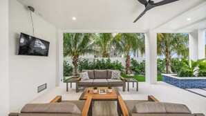 Un salon moderne comprenant un canapé, une table basse et des fauteuils, un téléviseur à écran plat fixé au mur, et une vue sur un jardin tropical avec des palmiers.