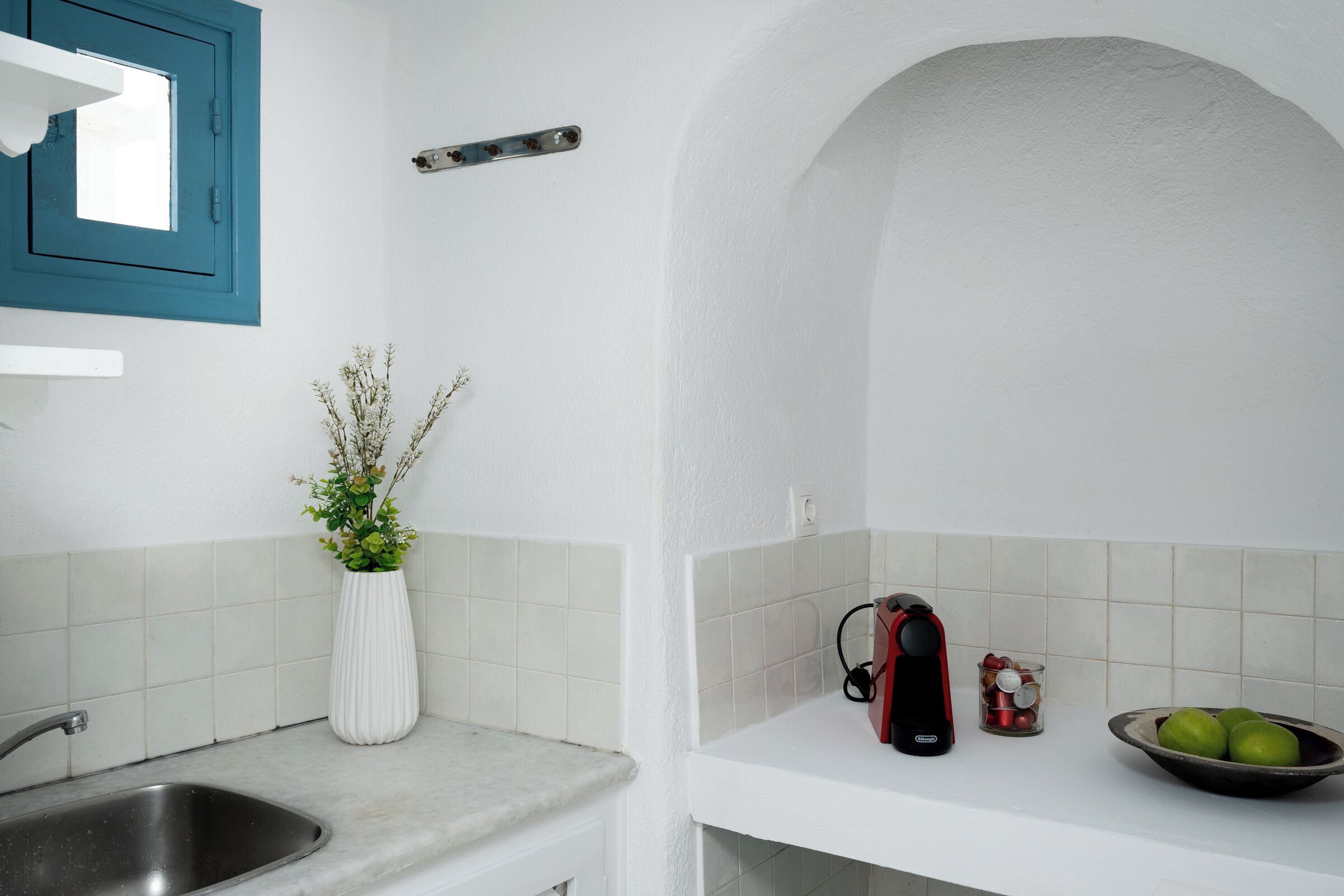 A kitchen with a sink, a white vase with plants, a kettle, and a bowl of fruit on the counter.
