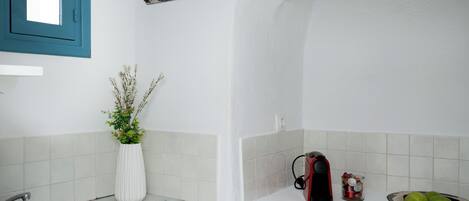 Una cocina con un lavamanos, un jarrón blanco con plantas, una tetera y un cuenco de fruta sobre la encimera.