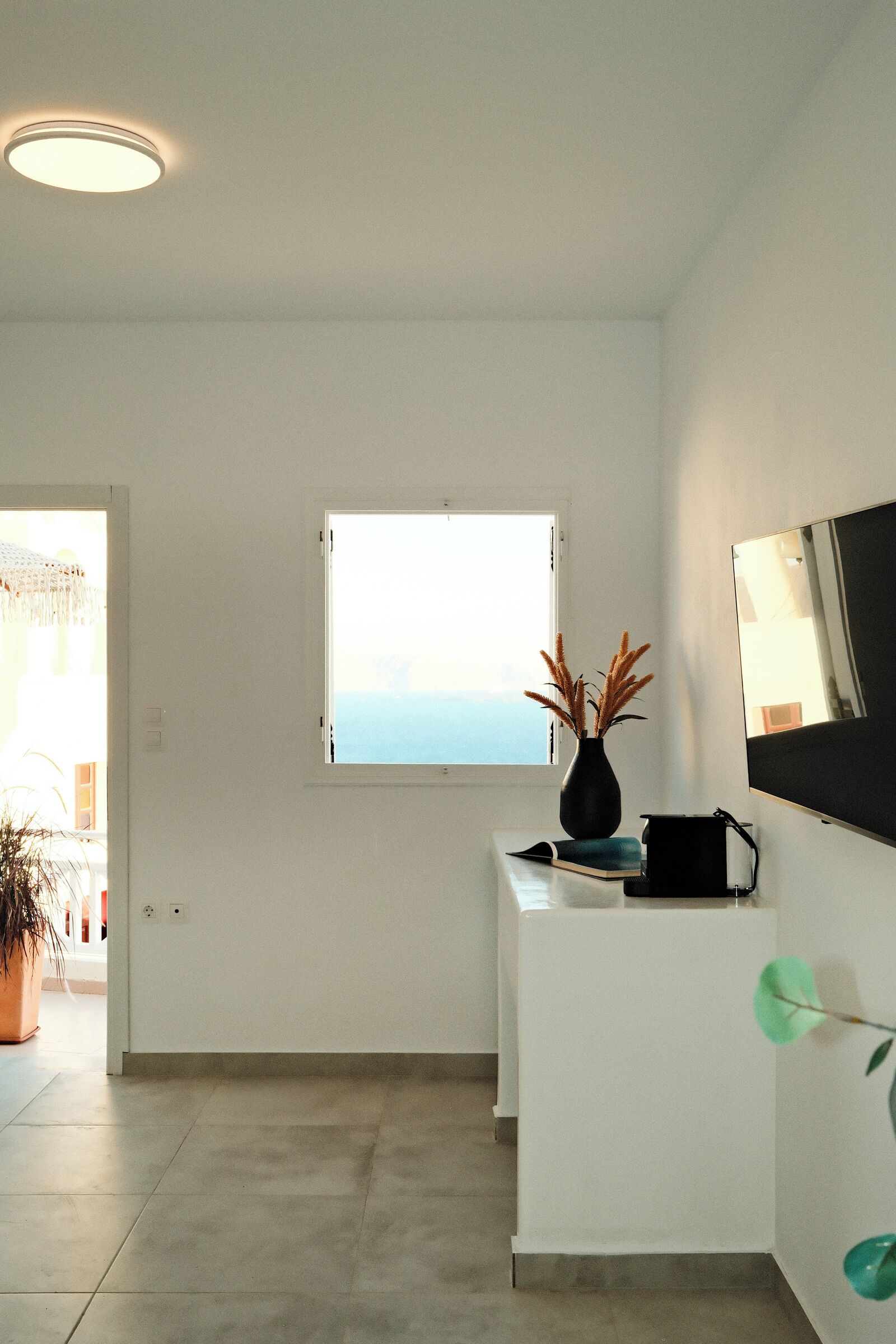A minimalist room with a white desk, a potted plant, a window with a view, and a flat-screen TV mounted on the wall.