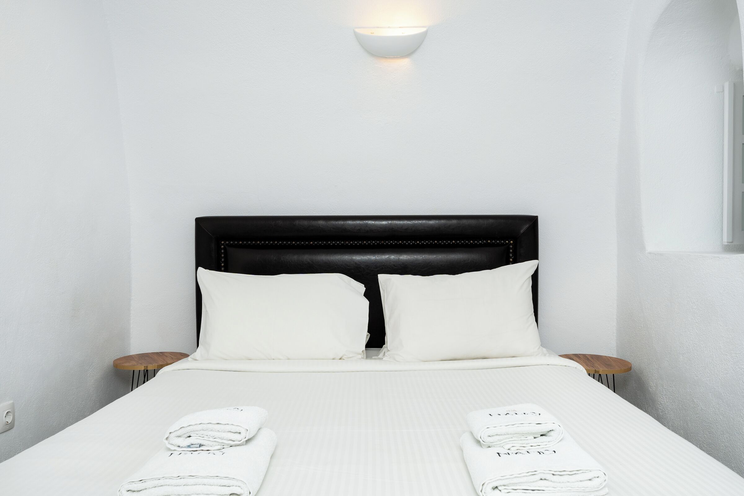 A neatly made bed with white linens, two pillows, and a black headboard. There are two bedside tables with lamps, and a wall-mounted light fixture above the bed.