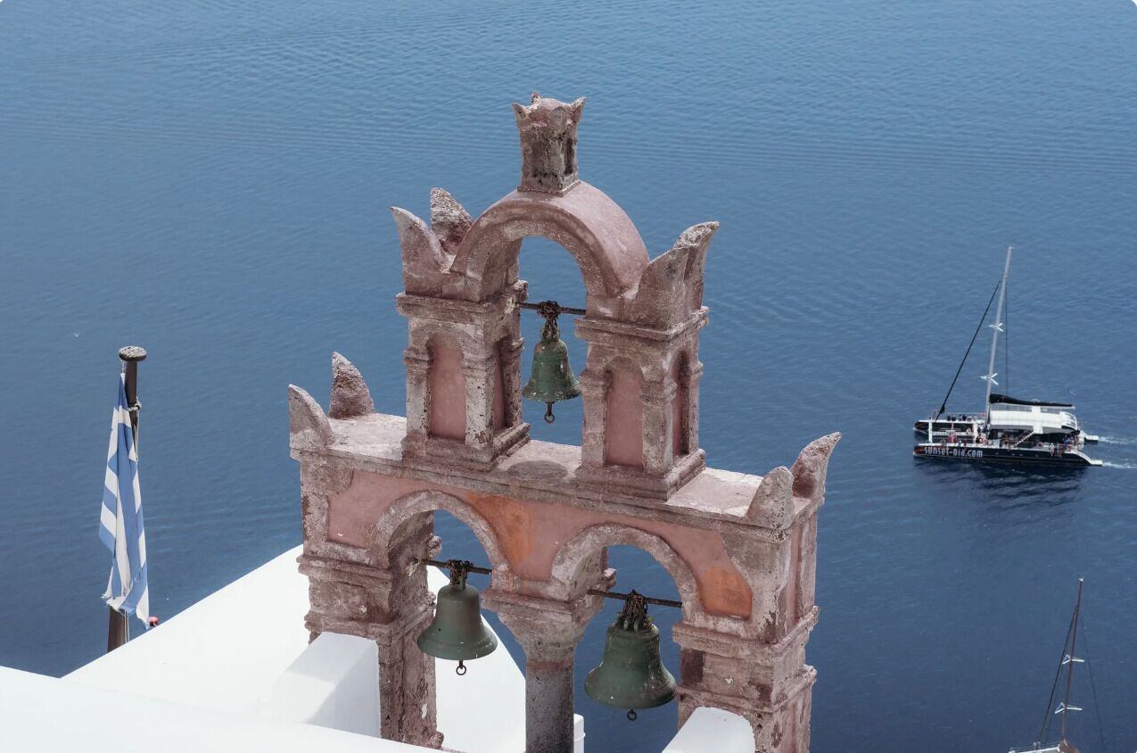 Un campanario con tres campanas, con vista a un mar tranquilo donde se divisa un barco a lo lejos.