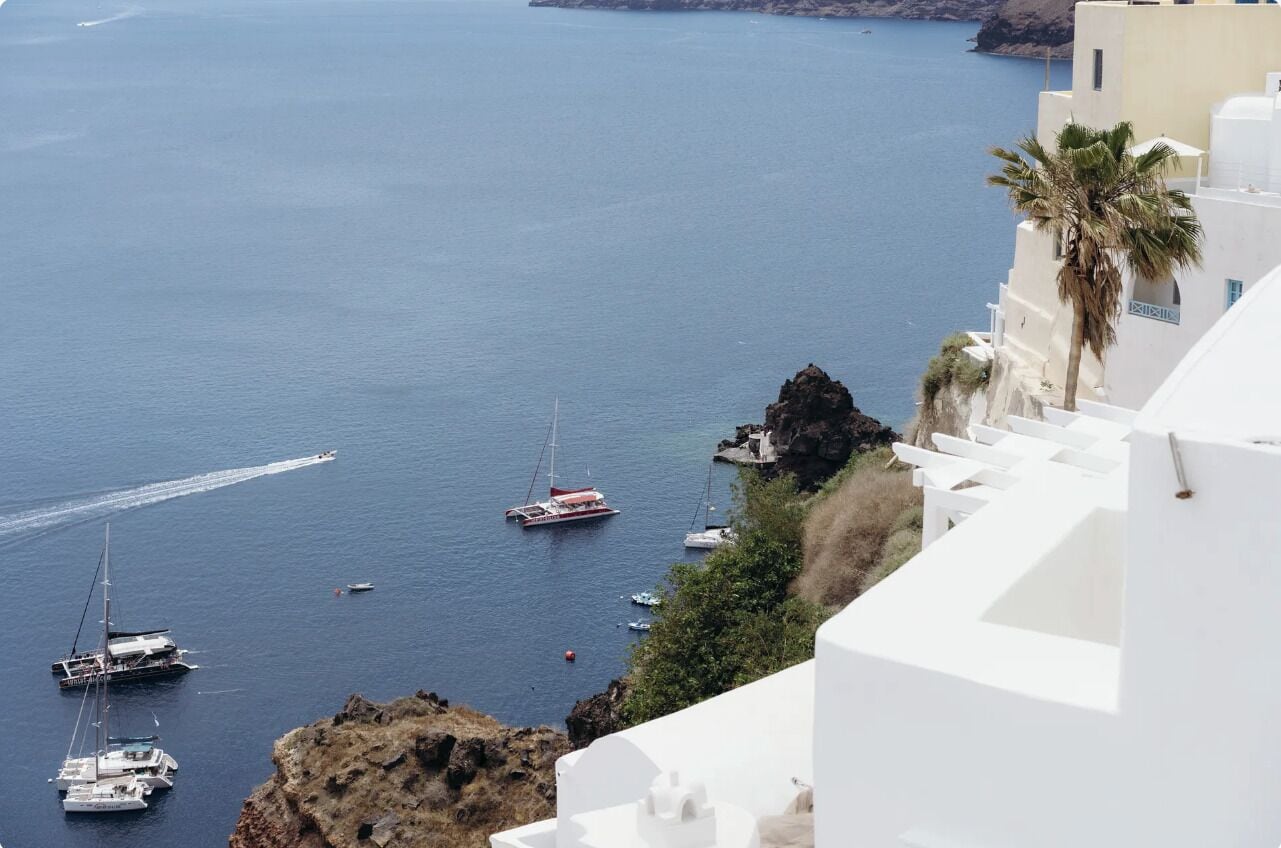 A coastal scene with white buildings, a rocky outcrop, and several boats on the water.