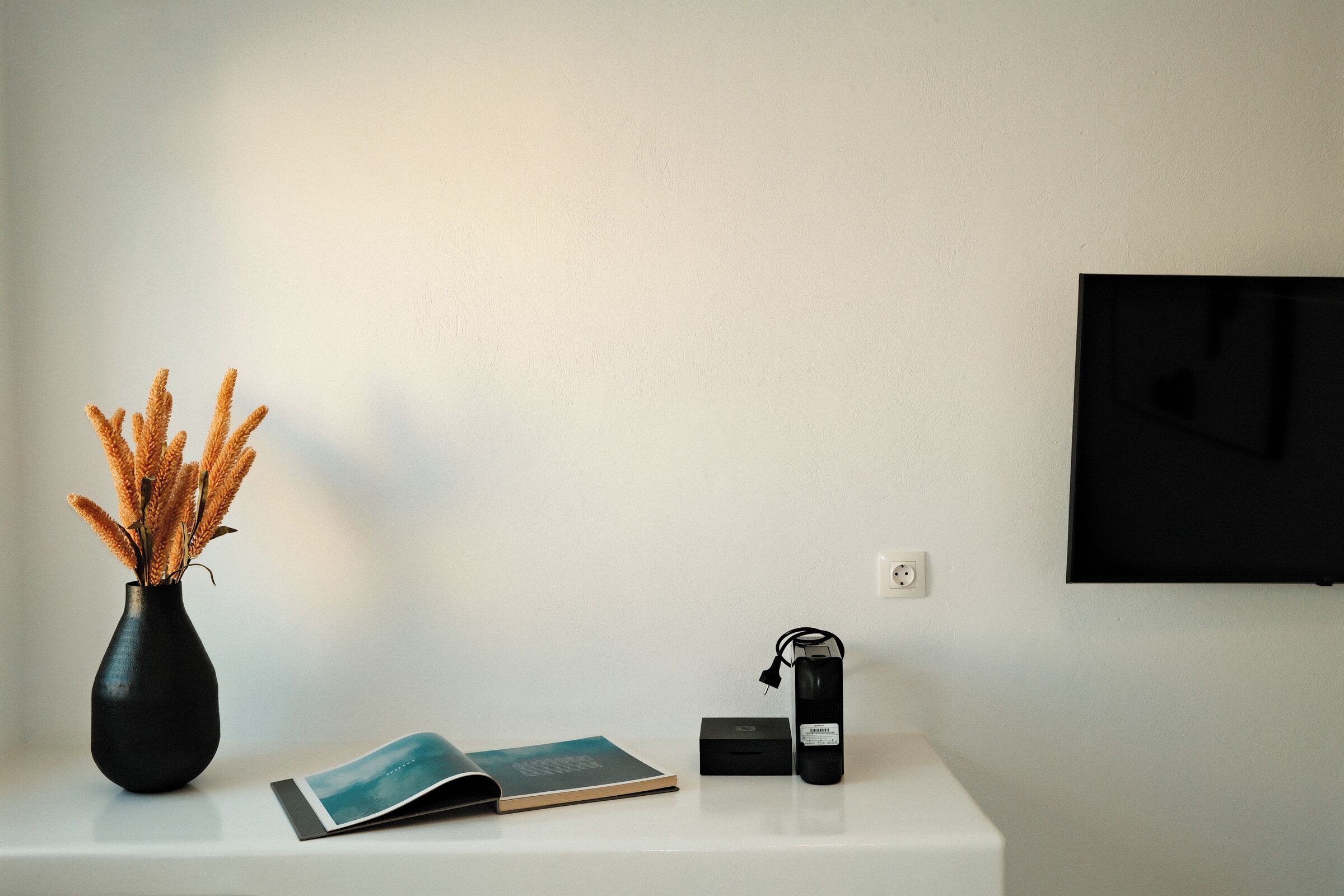 A white desk with a black vase of dried plants, a book, a black box, and a black electronic device.