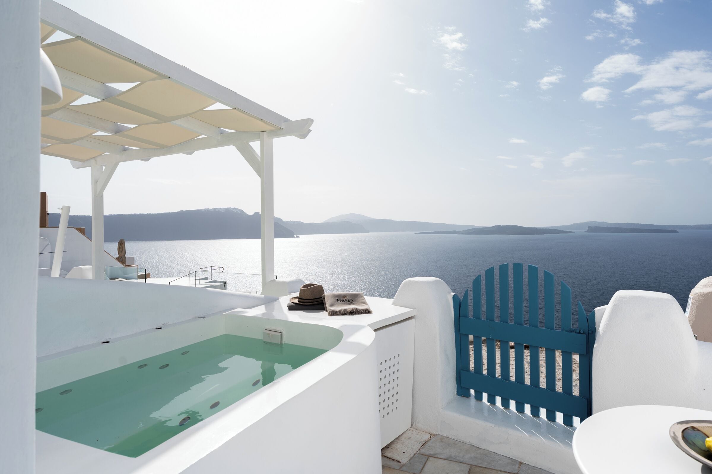 Un área de estar exterior blanca con piscina, una verja azul y vista al mar.