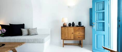 Ein schlicht eingerichteter Raum mit weißer Wand, blauer Tür, Holzschrank und grauem Sofa mit schwarzen Kissen.