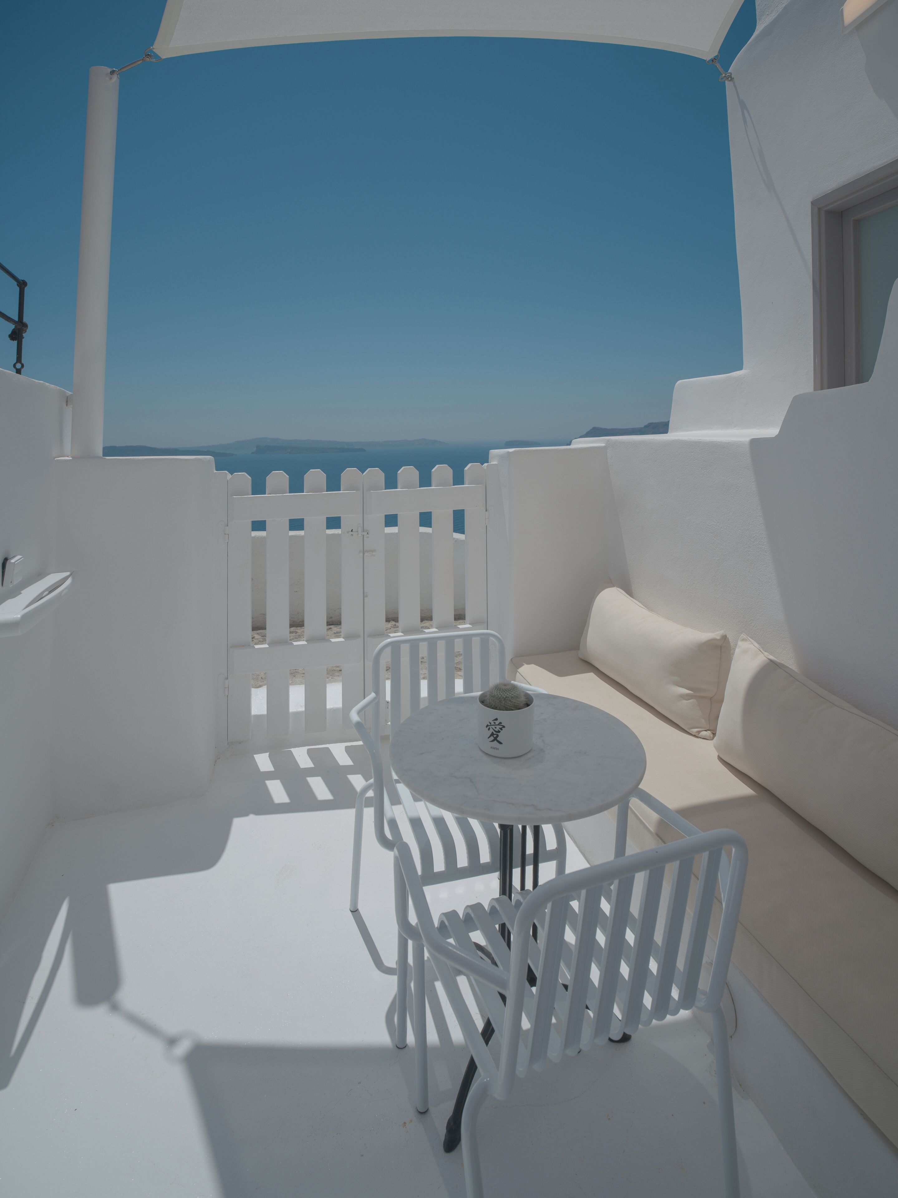 A white outdoor seating area with a table and chairs, a white fence, and a view of the sea.