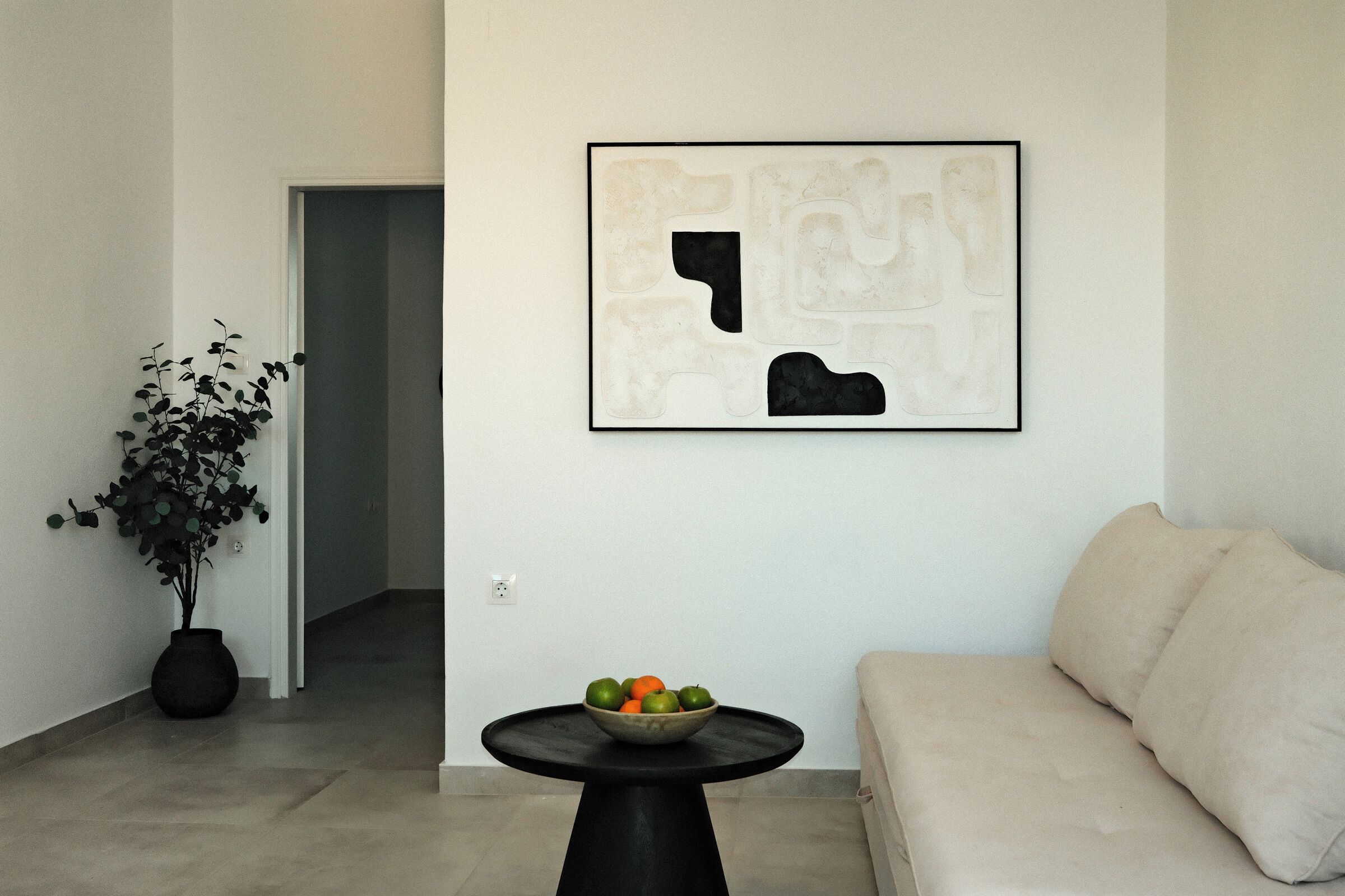 A modern living room with a sofa, a round black table with a bowl of fruit, a potted plant, and a framed abstract artwork on the wall.