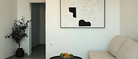 En moderne stue med en sofa, et rundt sort bord med en skål frugt, en potteplante og et indrammet abstrakt kunstværk på væggen.