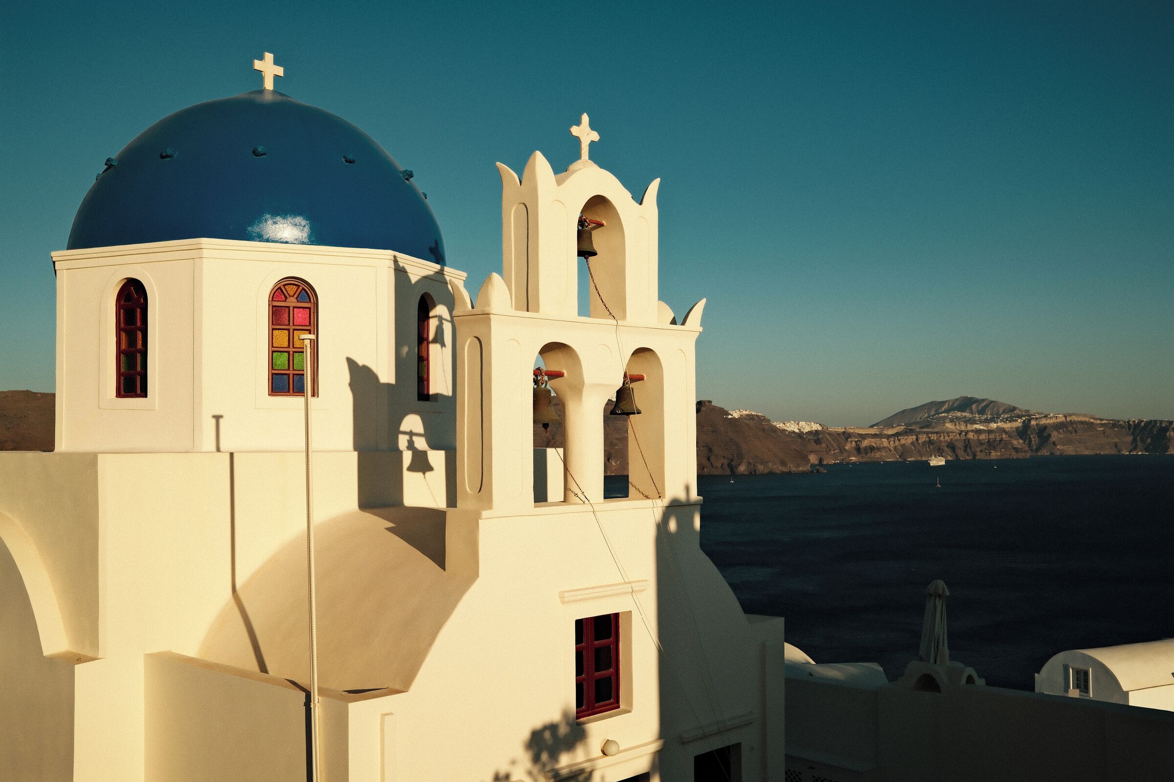 A white building with a blue dome and a bell tower, overlooking a body of water and mountains.